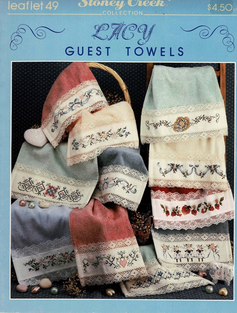 May include: A collection of six embroidered guest towels with lace trim. The towels are in various colors, including pink, blue, and cream. The embroidery designs include flowers, hearts, and a sheep. The towels are displayed on a wooden rack. The title "Lacy Guest Towels" is printed on the top of the image.