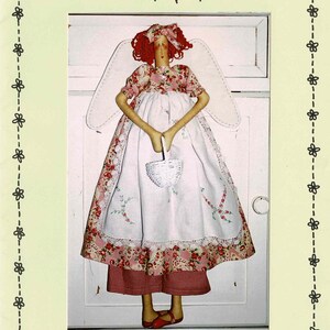 May include: A fabric doll named Charlotte by Rosalie Quinlan Designs with red hair, white wings, and a floral dress featuring a white apron and a small white basket. The doll wears red shoes and has a red hair accessory, presented on a pale yellow card.