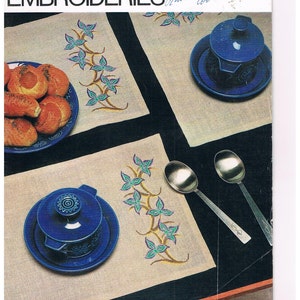 May include: A vintage sewing book cover, "COUNTED THREAD EMBROIDERIES", displays embroidered placemats with a floral pattern. The image includes blue ceramic dishes, spoons, and a plate of rolls.