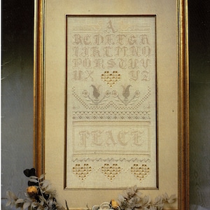 May include: Framed cross-stitch sampler with the text "The Peace Sampler" at the top. The sampler features an alphabet, decorative elements, and the word "PEACE". The frame is gold-toned, and the piece is decorated with dried flowers.
