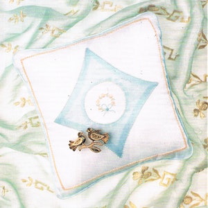 May include: A white pillow with a blue diamond shape in the center. The diamond has a circle with a floral design in the center. Two gold bird figurines are on the pillow.