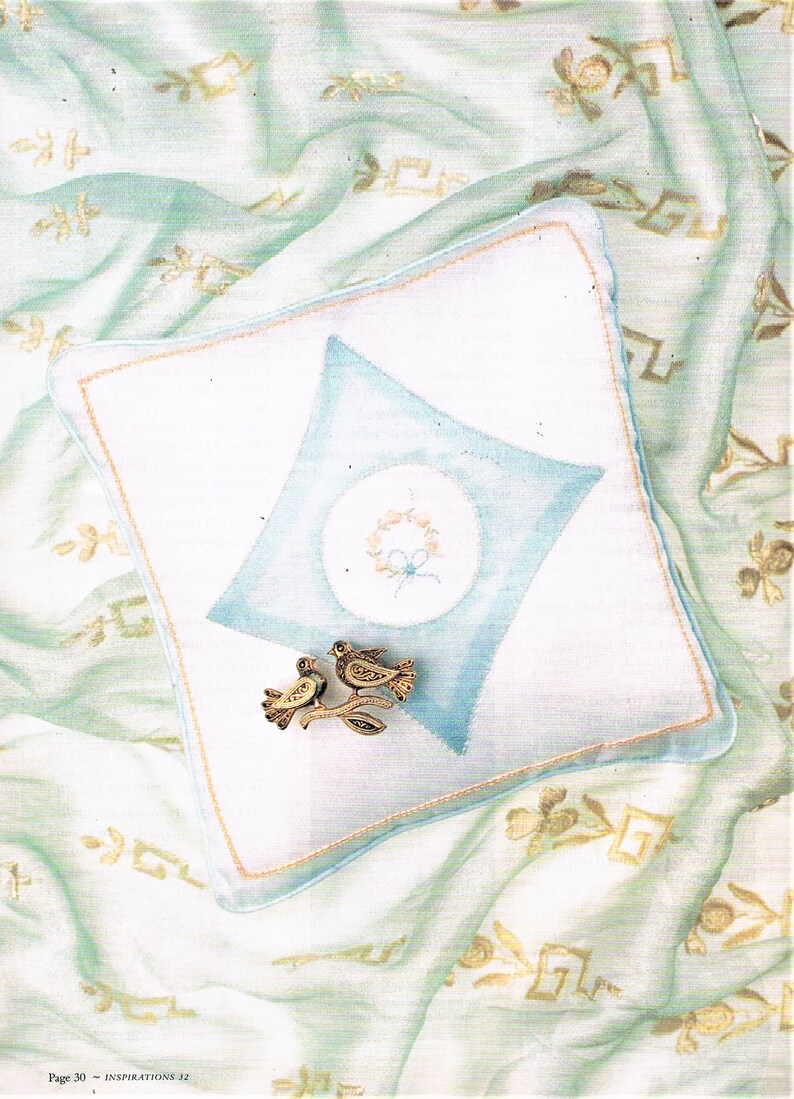 May include: A white pillow with a blue diamond shape in the center. The diamond has a circle with a floral design in the center. Two gold bird figurines are on the pillow.