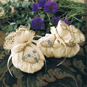 May include: Two cream fabric sachets, each adorned with embroidered blue flowers and gold trim, tied with white ribbons. The background shows purple flowers and green leaves.