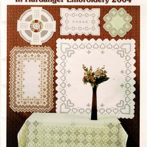 May include: A book cover titled Award-Winning Designs In Hardanger Embroidery 2004, compiled by Susan L. Meier and Rosalyn K. Watnemo, featuring various white Hardanger embroidery patterns on linens, including a tablecloth and wall hangings.