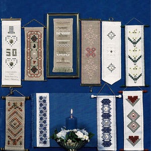 May include: A book cover titled Nordic Hardanger Bands III by members of the Nordic Needle Staff, featuring ten vertical fabric wall hangings with various embroidered patterns, a blue candle, and a floral arrangement on a deep blue background.