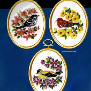 May include: Three oval-shaped embroidery hoops with gold frames. Each hoop features a different bird embroidered in colourful thread on white fabric. The first hoop shows a black and white bird with pink flowers. The second hoop shows a red bird with yellow flowers. The third hoop shows a yellow bird with purple flowers. Designs © Eleanor Engel.