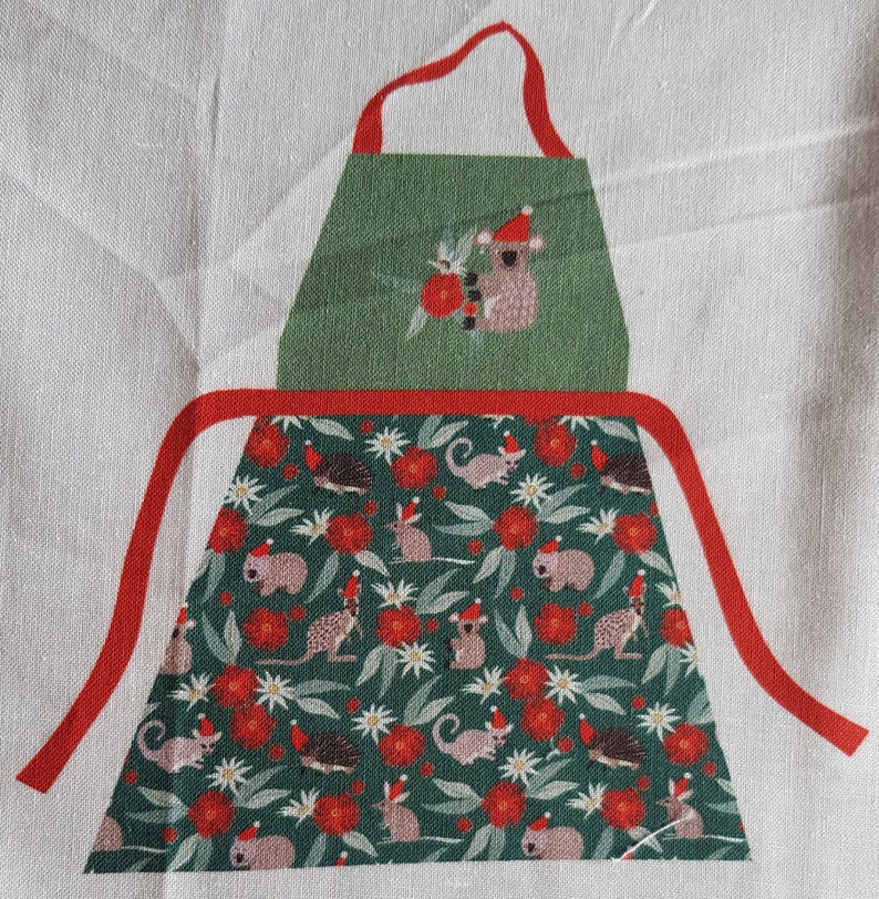 May include: Green and red Christmas-themed apron with a koala wearing a Santa hat on the top pocket. The apron has a repeating pattern of koalas, echidnas, and gum leaves.