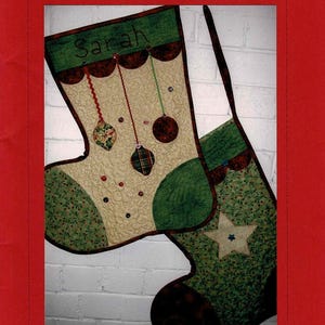 May include: Two Christmas stockings with a quilted design. One stocking has the name "Sarah" embroidered on it. The stockings are decorated with green, red, and brown accents, including ornaments and a star.