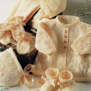 May include: A cream-colored baby clothing set, including a cardigan, bonnet, socks, mittens, and booties. The knitted pieces feature a delicate leaf pattern and are adorned with satin ribbons. The set is displayed on a white surface.