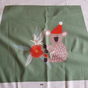 May include: A green fabric panel with a white koala wearing a red Santa hat and holding a red flower. The koala is surrounded by green leaves and white flowers. The fabric panel has the word "BIB" printed on the bottom.