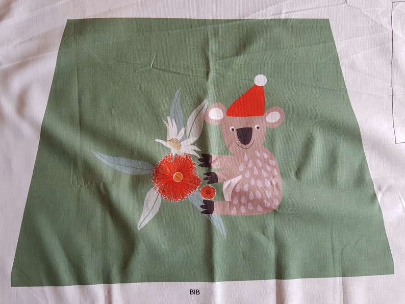 May include: A green fabric panel with a white koala wearing a red Santa hat and holding a red flower. The koala is surrounded by green leaves and white flowers. The fabric panel has the word "BIB" printed on the bottom.