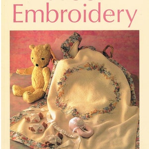 May include: The book cover reads "Wool Embroidery" with a soft pink background. The image shows a yellow teddy bear, a cream blanket with floral embroidery, and a baby rattle. The blanket is inside a basket with floral fabric.