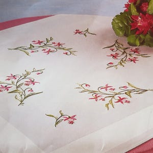 May include: White tablecloth with a delicate floral embroidery pattern featuring pink flowers and green leaves. The tablecloth has a pink border.