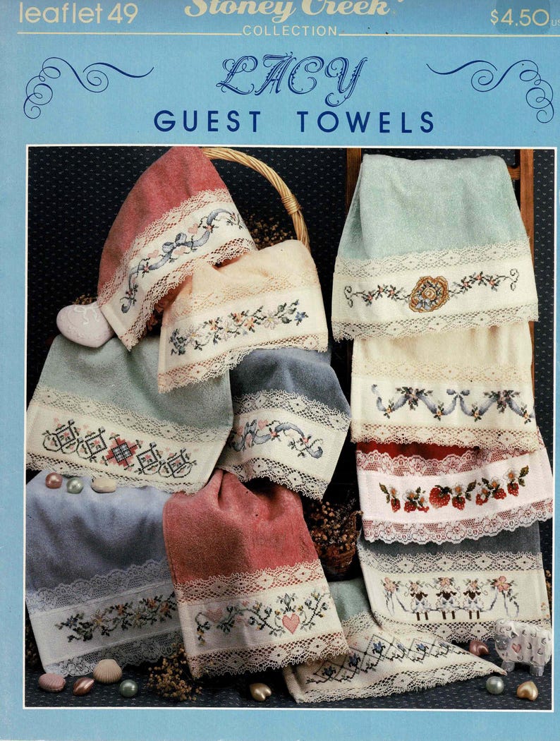 May include: A collection of six embroidered guest towels with lace trim. The towels are in various colors, including pink, blue, and cream. The embroidery designs include flowers, hearts, and a sheep. The towels are displayed on a wooden rack. The title "Lacy Guest Towels" is printed on the top of the image.