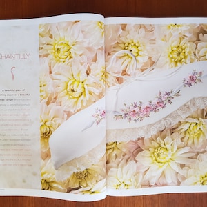 Puede incluir: Una percha blanca con bordados florales y ribete de encaje, exhibida en una p&aacute;gina con la palabra "CHANTILLY" y rodeada de flores color crema. La percha tiene un lazo de cinta.