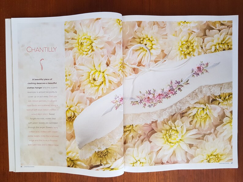 Puede incluir: Una percha blanca con bordados florales y ribete de encaje, exhibida en una p&aacute;gina con la palabra "CHANTILLY" y rodeada de flores color crema. La percha tiene un lazo de cinta.