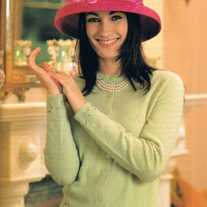 May include: A woman wearing a light green cardigan with pearl embroidery and a pink hat with pink flowers. She is wearing a pearl necklace and has dark hair.