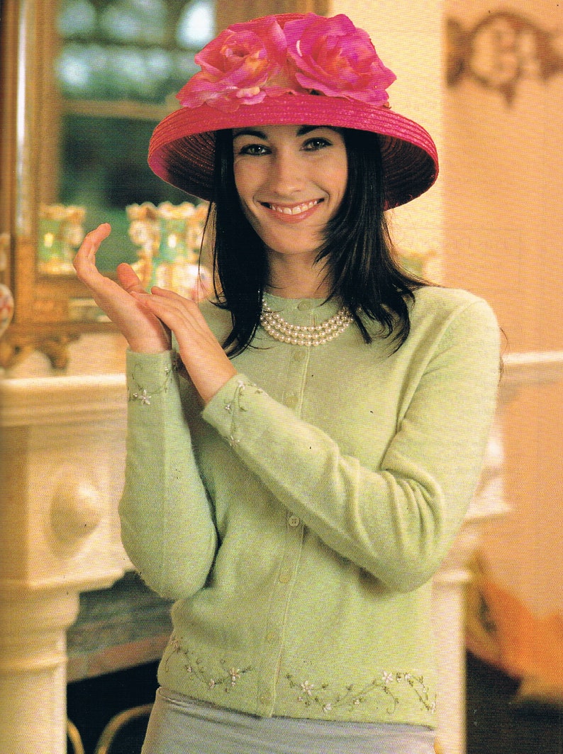 May include: A woman wearing a light green cardigan with pearl embroidery and a pink hat with pink flowers. She is wearing a pearl necklace and has dark hair.