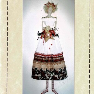 May include: A handmade doll wearing a white dress with red and white striped trim and a floral print skirt. The doll is holding a cushion with a star and the word "Joy" on it. The doll is wearing red and white striped stockings and red shoes.