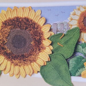 Puede incluir: Obra de arte bordada con girasoles. Los girasoles tienen p&eacute;talos amarillos y centros marrones. Hojas verdes y un fondo de cielo azul con una valla y p&aacute;jaros completan la escena. La obra de arte est&aacute; enmarcada en un borde blanco.