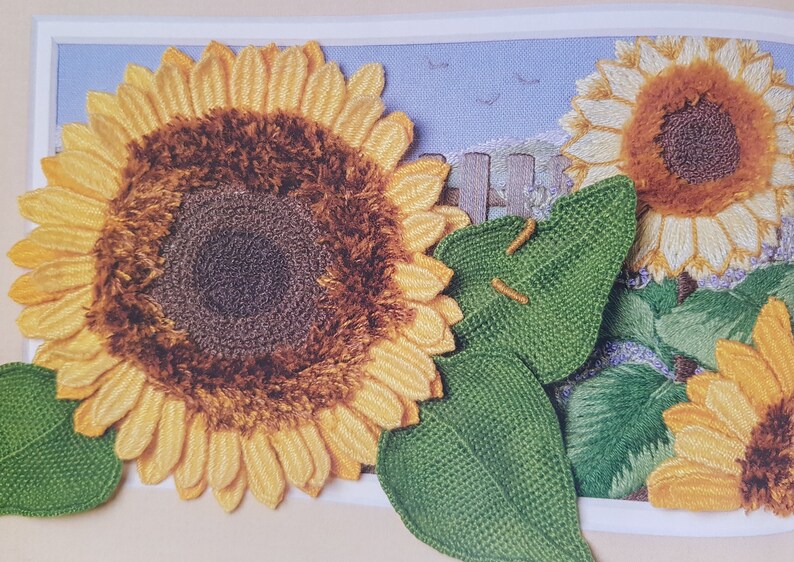 Puede incluir: Obra de arte bordada con girasoles. Los girasoles tienen p&eacute;talos amarillos y centros marrones. Hojas verdes y un fondo de cielo azul con una valla y p&aacute;jaros completan la escena. La obra de arte est&aacute; enmarcada en un borde blanco.