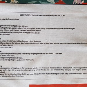 May include: A close-up of a white fabric with black text outlining instructions for sewing a Christmas apron. The instructions include steps for sewing the skirt, waist band, apron ties, and finishing touches. The text includes measurements in centimeters.