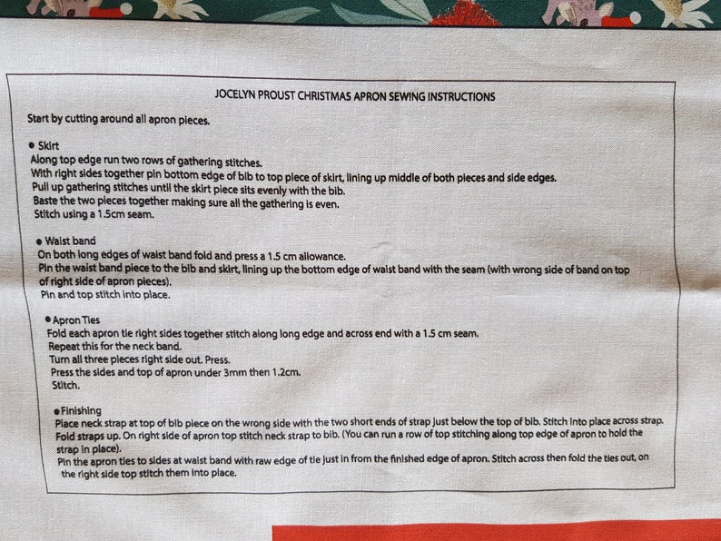 May include: A close-up of a white fabric with black text outlining instructions for sewing a Christmas apron. The instructions include steps for sewing the skirt, waist band, apron ties, and finishing touches. The text includes measurements in centimeters.