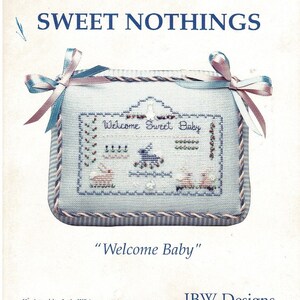 May include: A cross-stitch pattern for a baby sampler with a blue and pink ribbon border. The sampler features a house with the text "Welcome Sweet Baby" and a small scene with animals. The pattern is titled "Welcome Baby" and is from JBW Designs.