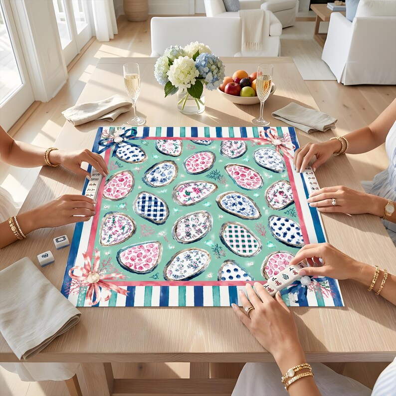 May include: A square mahjong mat featuring a light green background with a repeating pattern of oyster shells decorated in various blue, white, and pink floral and geometric prints, framed by a blue and white striped border with pink bows.