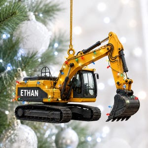 Op de afbeelding: Een geel met zwarte graafmachine-ornament hangend aan een gouden koord. Op de zijkant van de graafmachine staat de naam ETHAN in witte hoofdletters. Het bouwvoertuig is omwikkeld met kleine, kleurrijke, gloeiende kerstboomverlichting.
