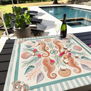 May include: A square, cream-colored game mat featuring a sea-themed design with seahorses, seashells, and coral in shades of orange, teal, and pink. The mat has a striped border and is adorned with game pieces. A bottle of champagne and a glass are on the table.
