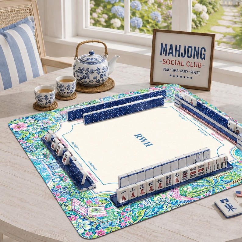 May include: A square mahjong mat with a colorful floral pattern, featuring a central cream area with the letters RWH. Blue and white mahjong tiles are arranged in racks on the mat. A blue and white teapot, two matching cups, and a sign reading MAHJONG SOCIAL CLUB are nearby.
