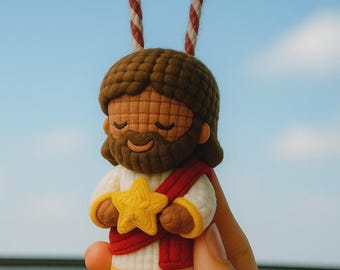 May include: A car charm featuring a plush figure of Jesus holding a yellow star. The figure has brown hair and beard, wearing a white robe with a red sash. The charm hangs from a striped red and white rope.