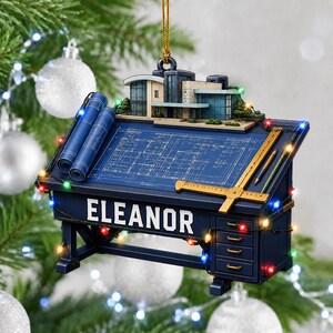 May include: A hanging ornament shaped like an architect's drafting table with the name ELEANOR printed on the front. The desk features a blue blueprint, two rolled blueprints, a T-square, a pencil, and a miniature modern house model on top.