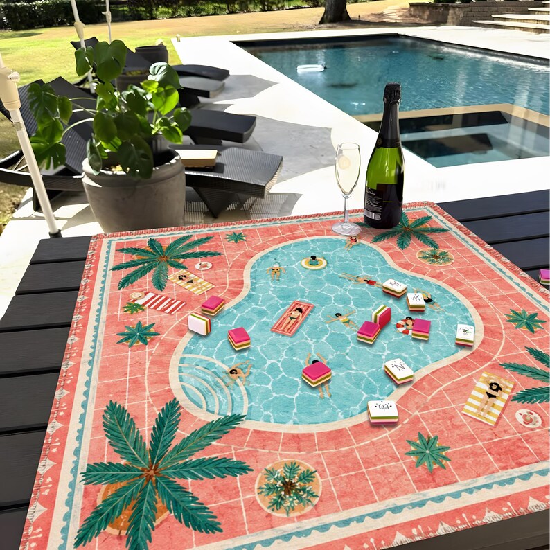 May include: A square game mat featuring a printed illustration of a swimming pool with people lounging on floats, surrounded by a coral-colored deck with palm trees. The mat is placed on a dark table with a bottle of champagne and a glass nearby.