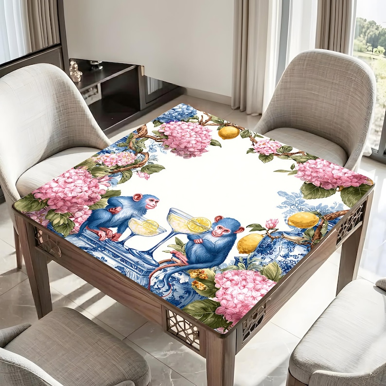 May include: Square wooden table with a decorative top featuring blue monkeys, pink hydrangeas, lemons, and blue and white porcelain patterns. The table has a light wood finish and is surrounded by chairs.