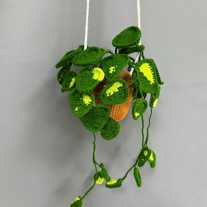 May include: A handmade crochet hanging plant with vibrant green leaves and yellow accents. The plant is in an orange pot and suspended by white cords against a gray background. The leaves have a detailed, textured appearance.