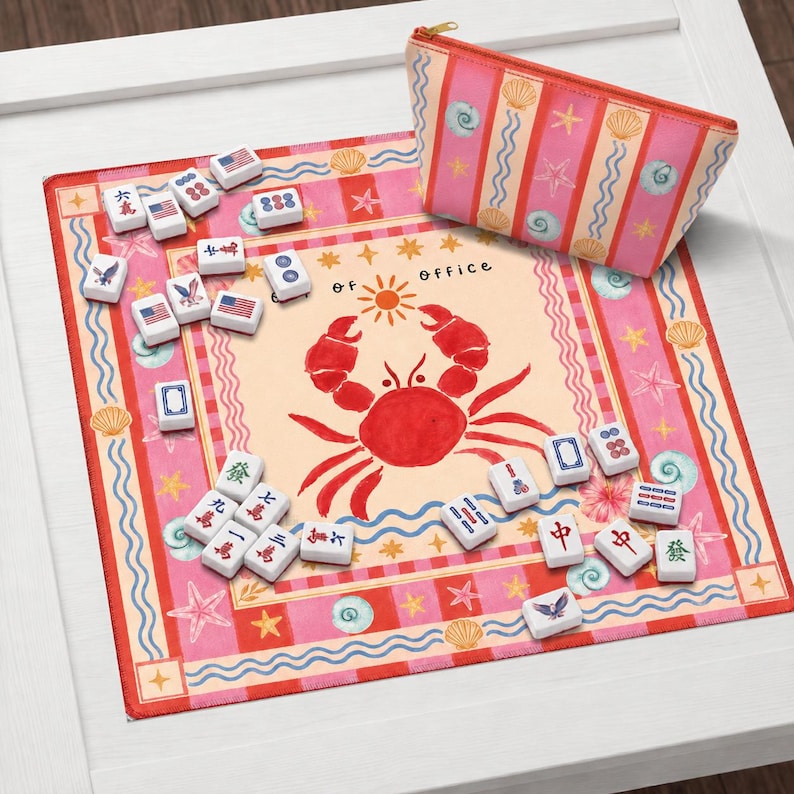 K&ouml;nnte beinhalten: Ein farbenfrohes Mahjong-Set mit Spielsteinen auf einer rosa-beige farbenen Spielmatte mit einer Krabbenillustration und dem Text "of office". Ein passendes Etui mit gestreiftem Muscheldesign ist ebenfalls vorhanden.