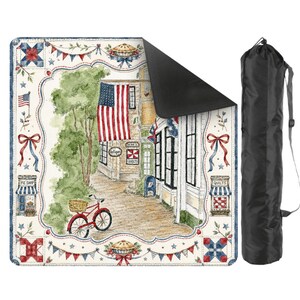 May include: A picnic blanket with a patriotic design, showing a watercolour illustration of a charming American town with an American flag, a red bicycle, and a pie shop. Includes a black carry bag.