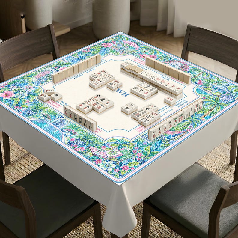 May include: A square mahjong table cover featuring a vibrant floral and botanical print in shades of blue, green, and pink. The center surface is cream-colored with printed game instructions, the letters WH, and several rows of mahjong tiles.