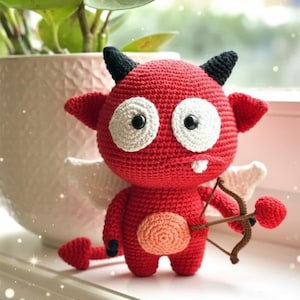 May include: A handmade red crochet devil doll with black horns, white eyes, and small white wings. The doll holds a brown bow and a red heart-shaped arrow. The doll has a peach-coloured belly and a heart-shaped tail.
