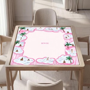 May include: A square table mat with a light pink center featuring the letters RWH in pink. The border consists of multiple irregular pink-rimmed pools filled with blue water, green palm trees, and small figures swimming or relaxing in the water.