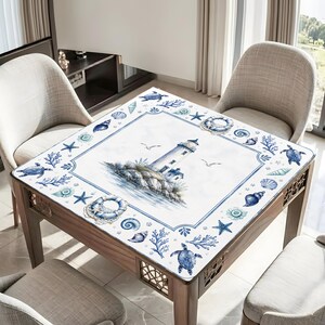 Puede incluir: Mesa cuadrada de madera con dise&ntilde;o n&aacute;utico. La superficie presenta una ilustraci&oacute;n de faro rodeada de conchas marinas, estrellas de mar y tortugas en tonos azules y blancos. La mesa tiene un marco de madera y est&aacute; rodeada de sillas.