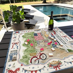 May include: A patriotic-themed table runner featuring a watercolour illustration of a small town, an American flag, and a red bicycle. The runner is decorated with red, white, and blue accents and a pie shop.