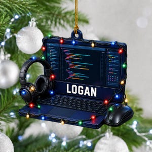 May include: A dark blue laptop-shaped Christmas ornament featuring a screen displaying lines of code, the name LOGAN in white text, a pair of black headphones, and a black computer mouse, all wrapped in a string of multicoloured holiday lights.