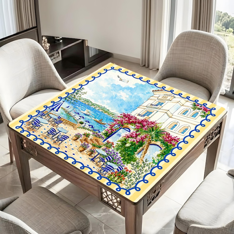 Puede incluir: Mesa cuadrada de madera con una tapa decorativa que presenta una escena costera. La obra de arte representa un caf&eacute; junto al mar con sombrillas, mesas y sillas azules y blancas. La mesa tiene un borde amarillo con un dise&ntilde;o de olas azules.