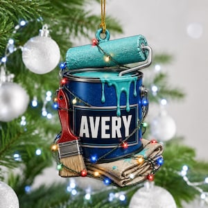 Puede incluir: Un adorno navideño colgante con forma de lata de pintura azul con el nombre AVERY impreso en blanco. Un rodillo de pintura turquesa descansa encima, con pintura goteando por el lateral. Un pincel de mango rojo y un paño doblado están debajo.
