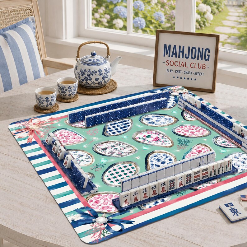 May include: A square mahjong mat with a teal background featuring oyster shell patterns, blue and white tile racks, and white mahjong tiles with red and black characters. A blue and white teapot, two matching teacups, and a sign reading MAHJONG SOCIAL CLUB are present.