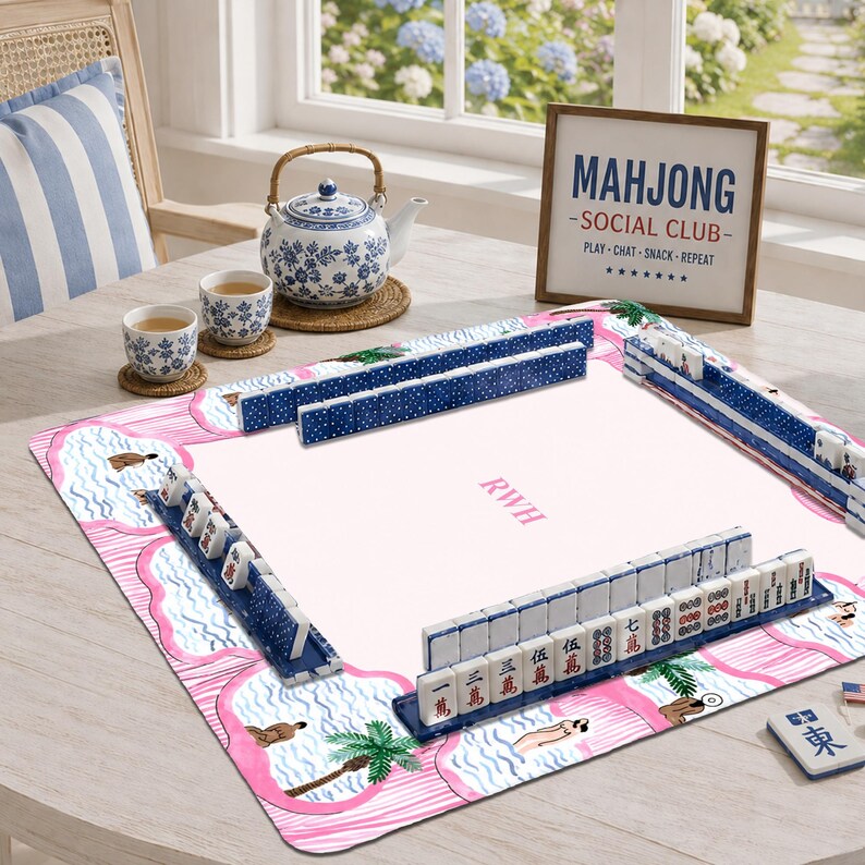May include: A mahjong set featuring a pink and white play mat with palm tree and water illustrations, blue tile racks, and white tiles with black and red characters. A white and blue floral teapot and two matching cups sit on the table.