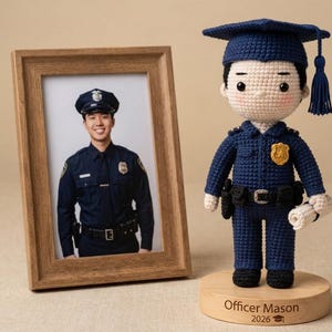 Op de afbeelding: Een ingelijste foto van een glimlachende politieagent staat naast een gehaakte pop in politie-uniform en afstudeerhoed. De pop staat op een houten voet met de naam "Officer Mason" en het jaar "2026" gegraveerd. De pop houdt een opgerold document vast.